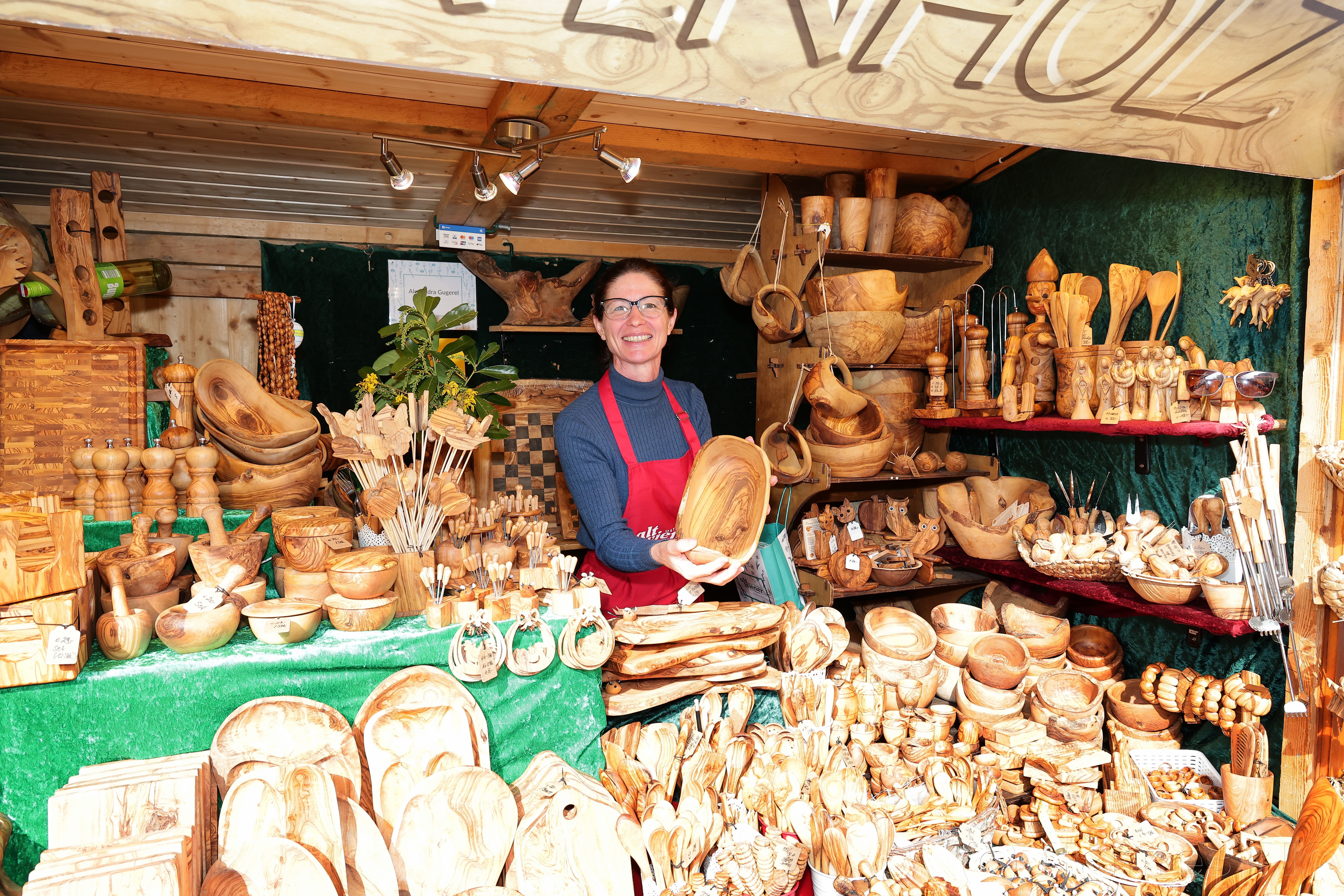 Woman in a red apron stands in a wooden crafts stall, displaying a carved bowl. Various wooden items fill the shelves around her.