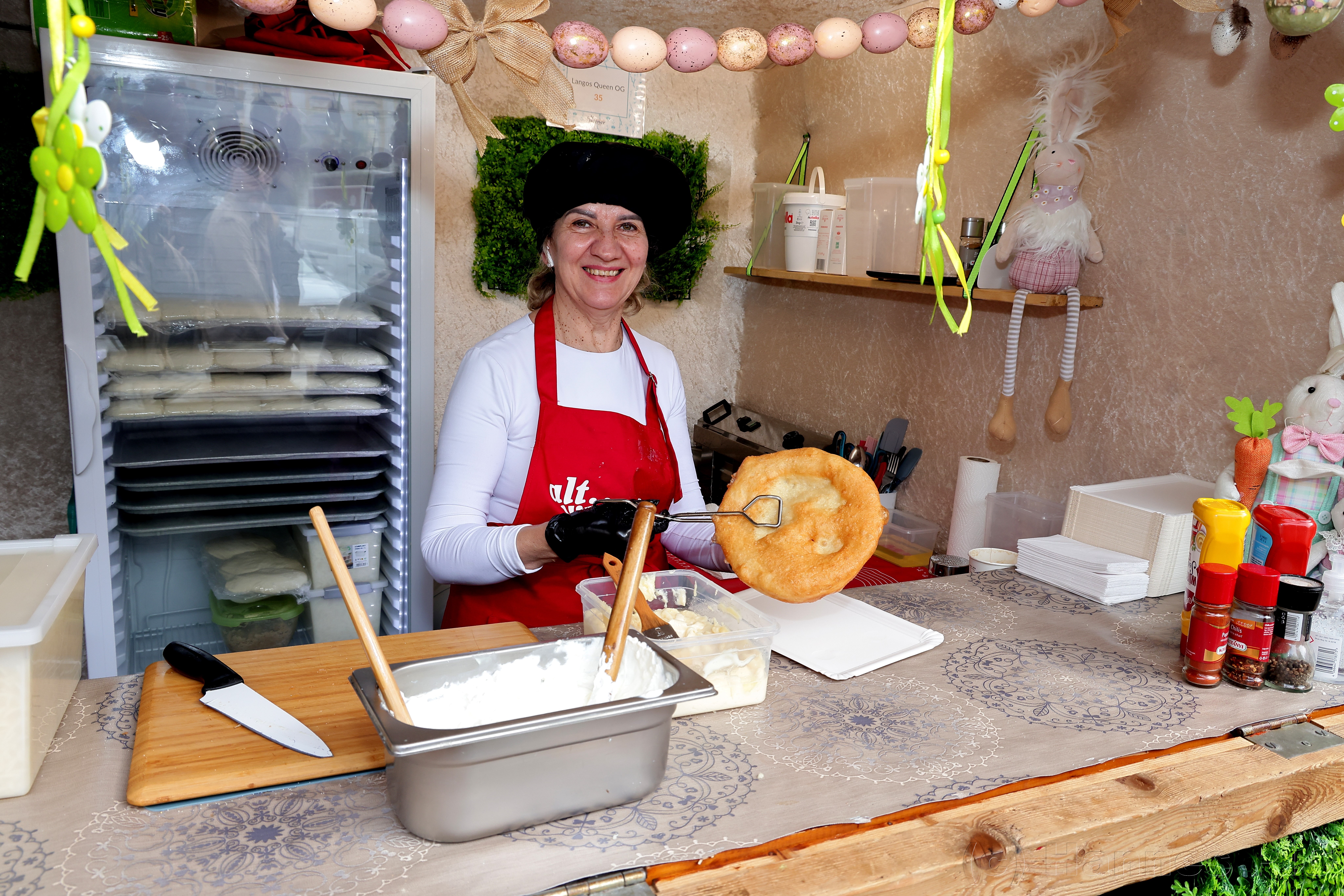 Smiling vendor in a red apron holds a langos at a decorated food stall, with cooking utensils and condiments displayed.