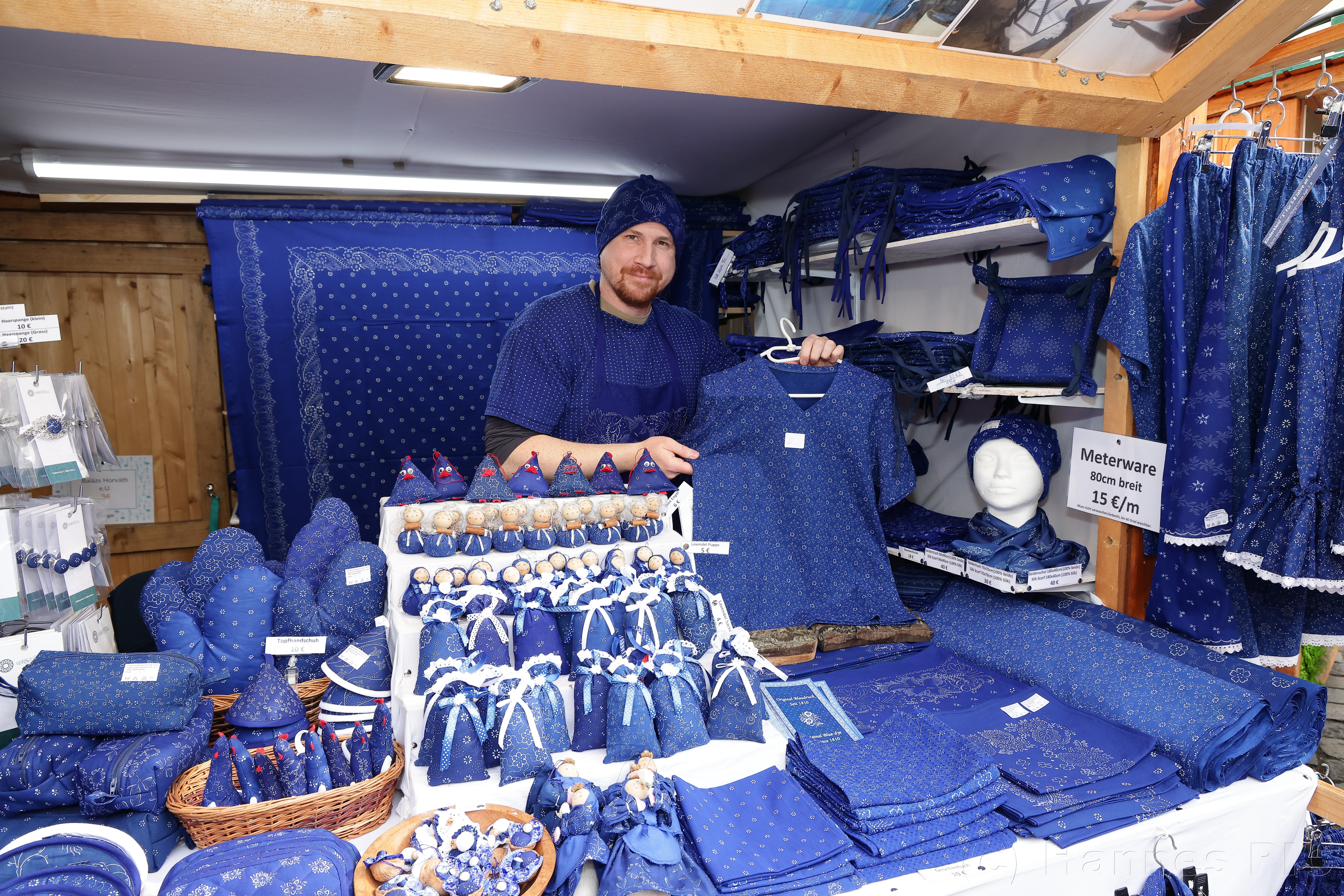 Man in a blue outfit showcasing blue textile goods at a market stall, surrounded by various fabric items and accessories.