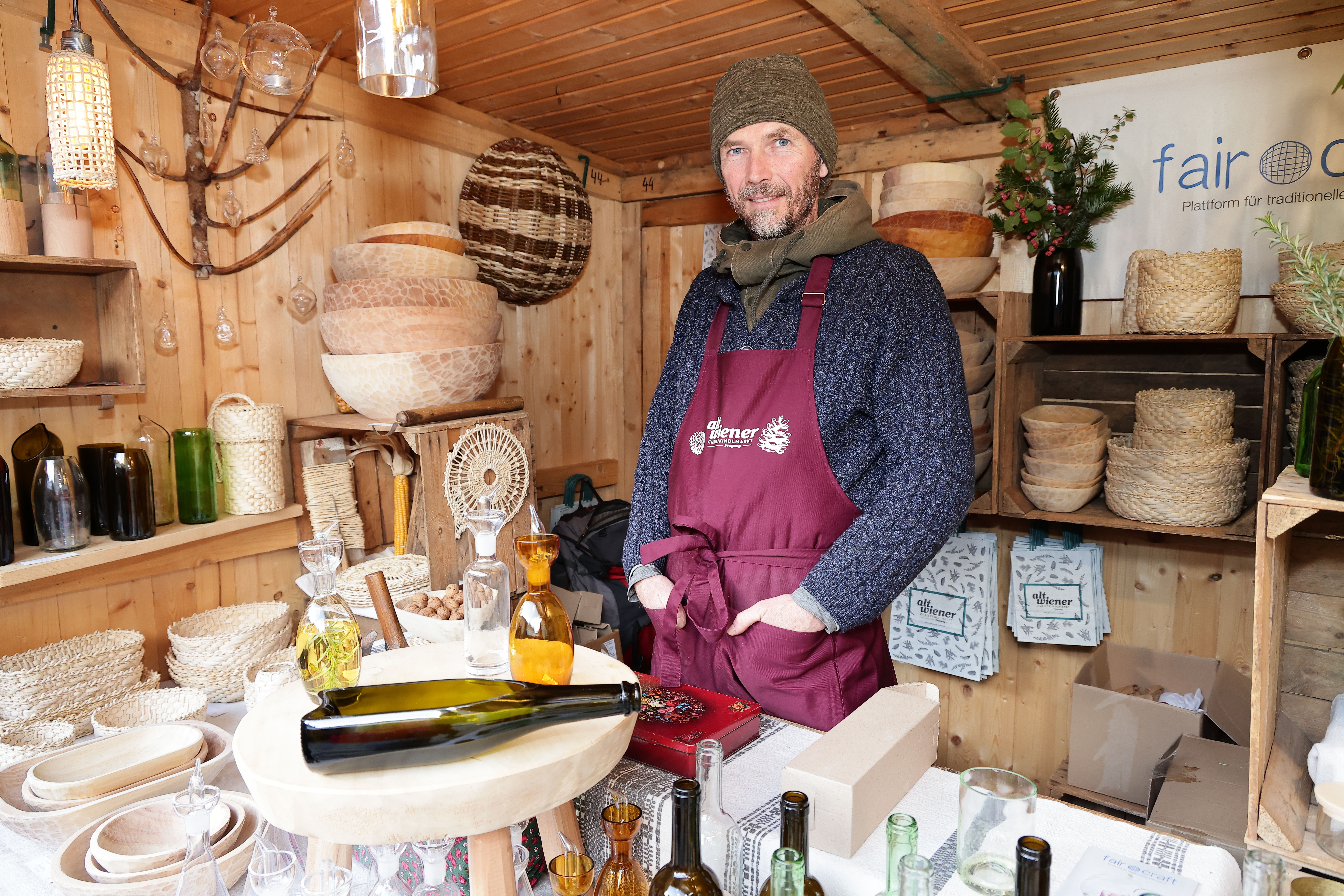 Man in a maroon apron stands in a wooden booth displaying handmade baskets, glass bottles, and crafts.