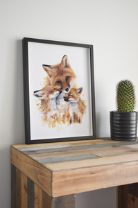 Framed fox illustration on a wooden table next to a small potted cactus against a white wall.