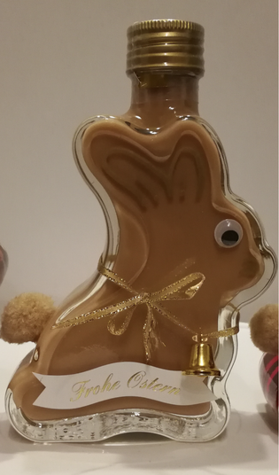Bunny-shaped bottle of caramel liqueur with a gold cap, gold ribbon, small bell, pom-pom tail, and "Frohe Ostern" label.