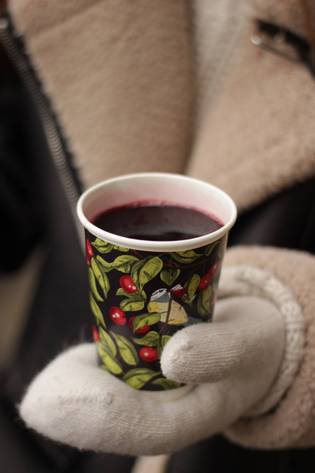 A person in a brown coat and white gloves holds a cup with a leafy, red berry pattern, filled with a dark beverage.
