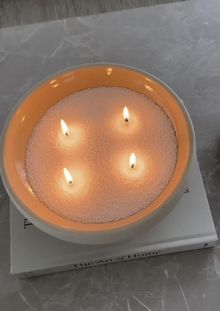 A lit candle with four wicks in a white bowl sits on a closed book titled "The Art of Home" on a gray marble surface.