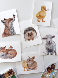 Watercolor paintings of a fox, hedgehog, duckling, lamb, and rabbit with flowers, arranged on a white surface with a paint palette nearby.