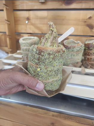 Hand holding a chimney cake covered in green sprinkles, topped with ice cream in a paper tray. Wooden background with more cakes in the back.