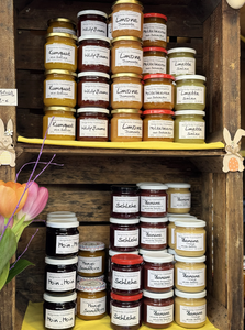 Jars of assorted fruit jams and marmalades stacked on wooden shelves, with decorative flowers and Easter ornaments.