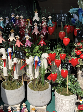 Decorative plant pots with ceramic stork figures, red heart and flower ornaments, and green foliage on display with price tags.