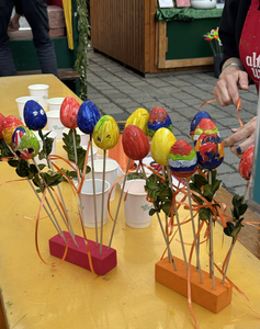 Colorful, decorated eggs on sticks with ribbons are arranged in wooden holders on a table, with cups and crafting supplies nearby.