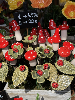 Ceramic mushrooms and flowers with ladybugs displayed for sale. A chalkboard sign shows prices: 1 for 4.50€, 5 for 20€.
