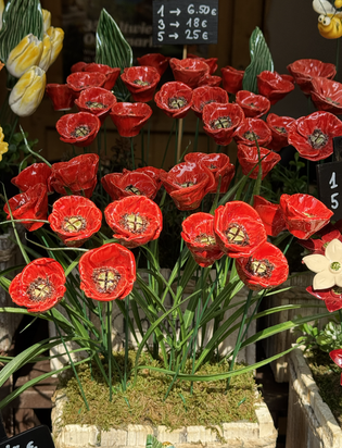Vibrant display of ceramic red poppies with green stems, set on a mossy base. Various price tags in Euros are visible in the background.