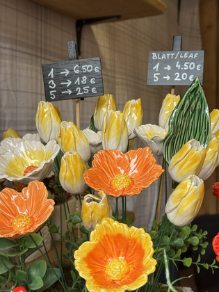 Colorful ceramic flowers with orange and yellow petals displayed at a market, accompanied by two price signs in the background.