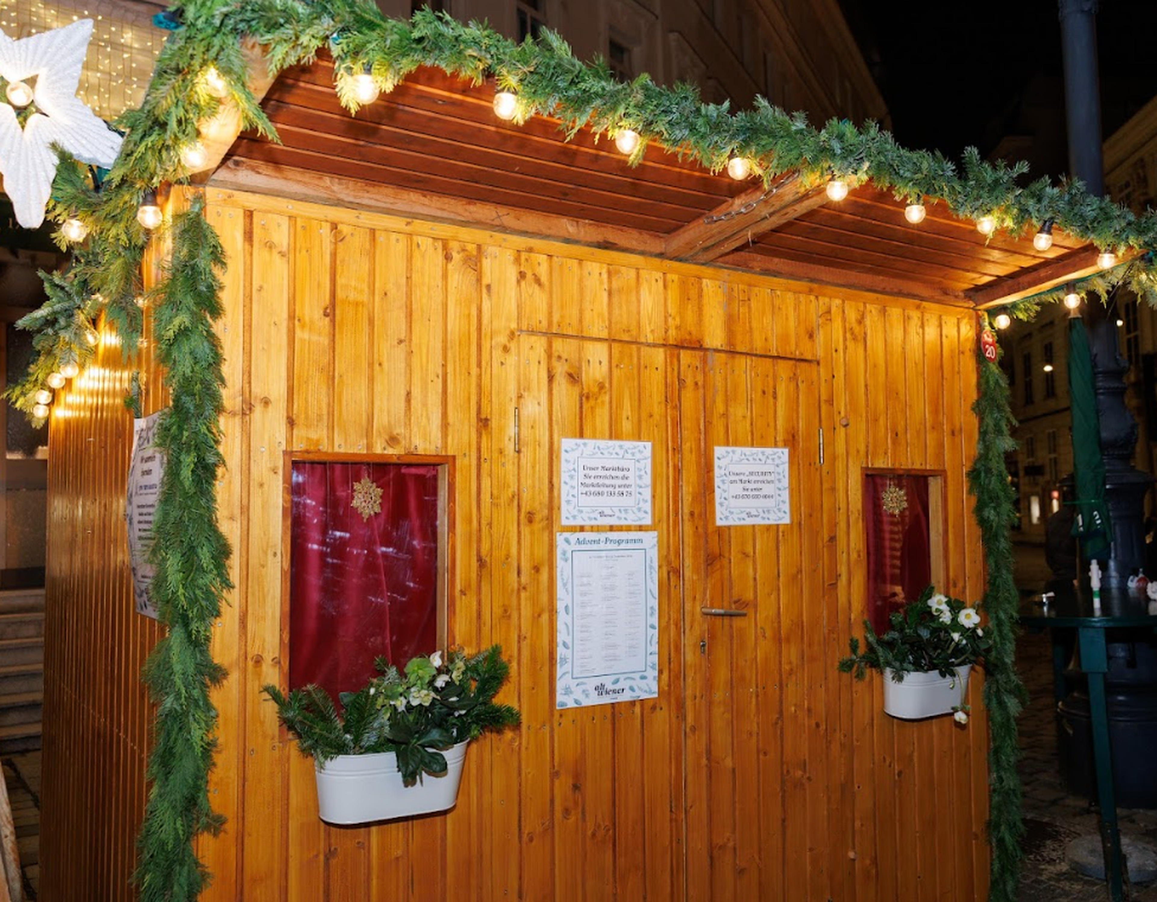 Cozy wooden Christmas market stall adorned with greenery and lights. Signs and floral decorations create a festive, inviting atmosphere at night.