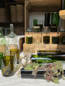 A display of glassware made from recycled green and brown glass bottles, set on wooden shelves with potted succulents nearby.