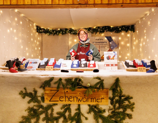 A smiling vendor at a festive stall sells colorful foot warmers. The cozy booth is adorned with string lights and pine garlands, creating a warm atmosphere.