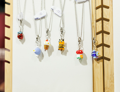 Close-up of six playful glass pendants on silver chains. Designs include a ladybug, cupcake, bee, and mushroom, displayed on a wooden rack. Bright and whimsical.