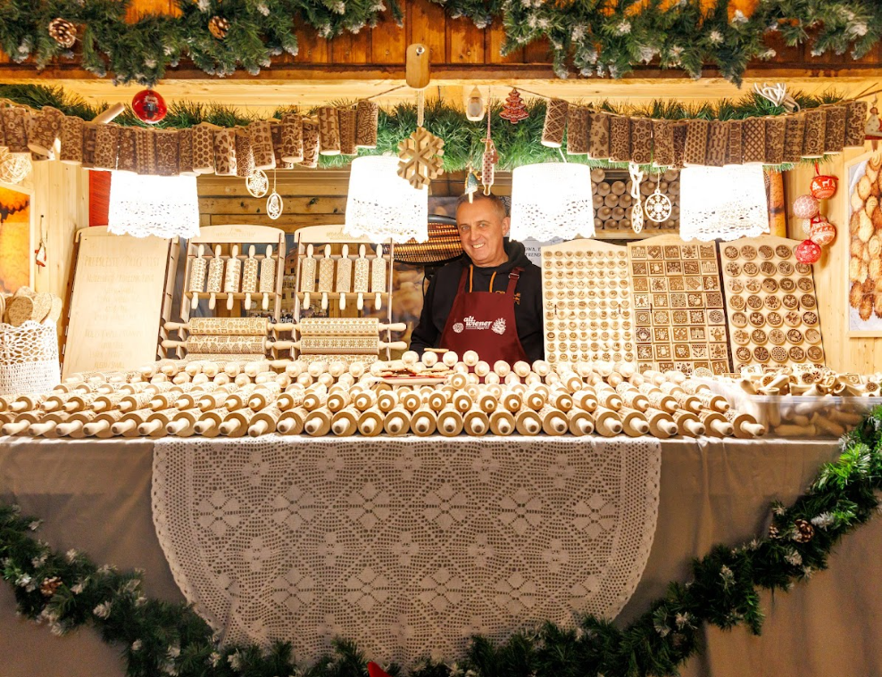 A vendor smiles behind a festive wooden stall at a Christmas market, displaying intricately patterned rolling pins. Green garlands and lights adorn the space.