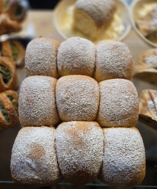 Freshly baked batch of nine soft rolls dusted with powdered sugar on a tray, surrounded by other pastries in the background.