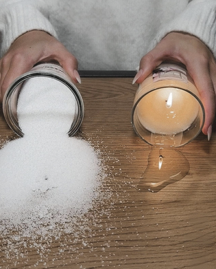 Hands tipping a jar of white powder and a lit candle, spilling wax, on a wooden surface.