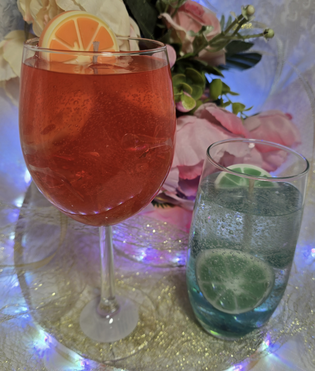 Two cocktails on a table: one red with a lemon slice, the other blue with a lime slice, surrounded by flowers and fairy lights.