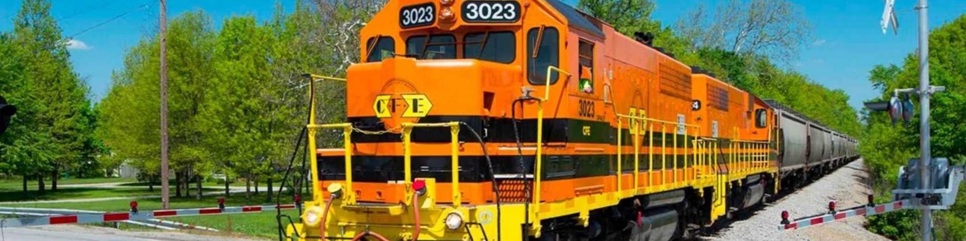 Bright orange freight train traveling through a rural crossing on a sunny day, with trees and a clear blue sky in the background.
