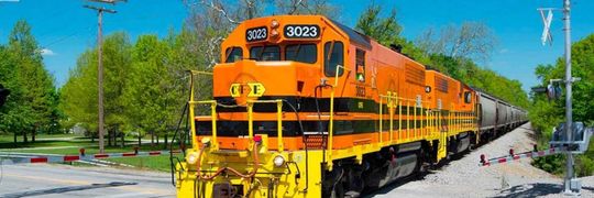 Bright orange freight train traveling through a rural crossing on a sunny day, with trees and a clear blue sky in the background.