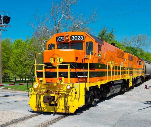 Bright orange freight train traveling through a rural crossing on a sunny day, with trees and a clear blue sky in the background.