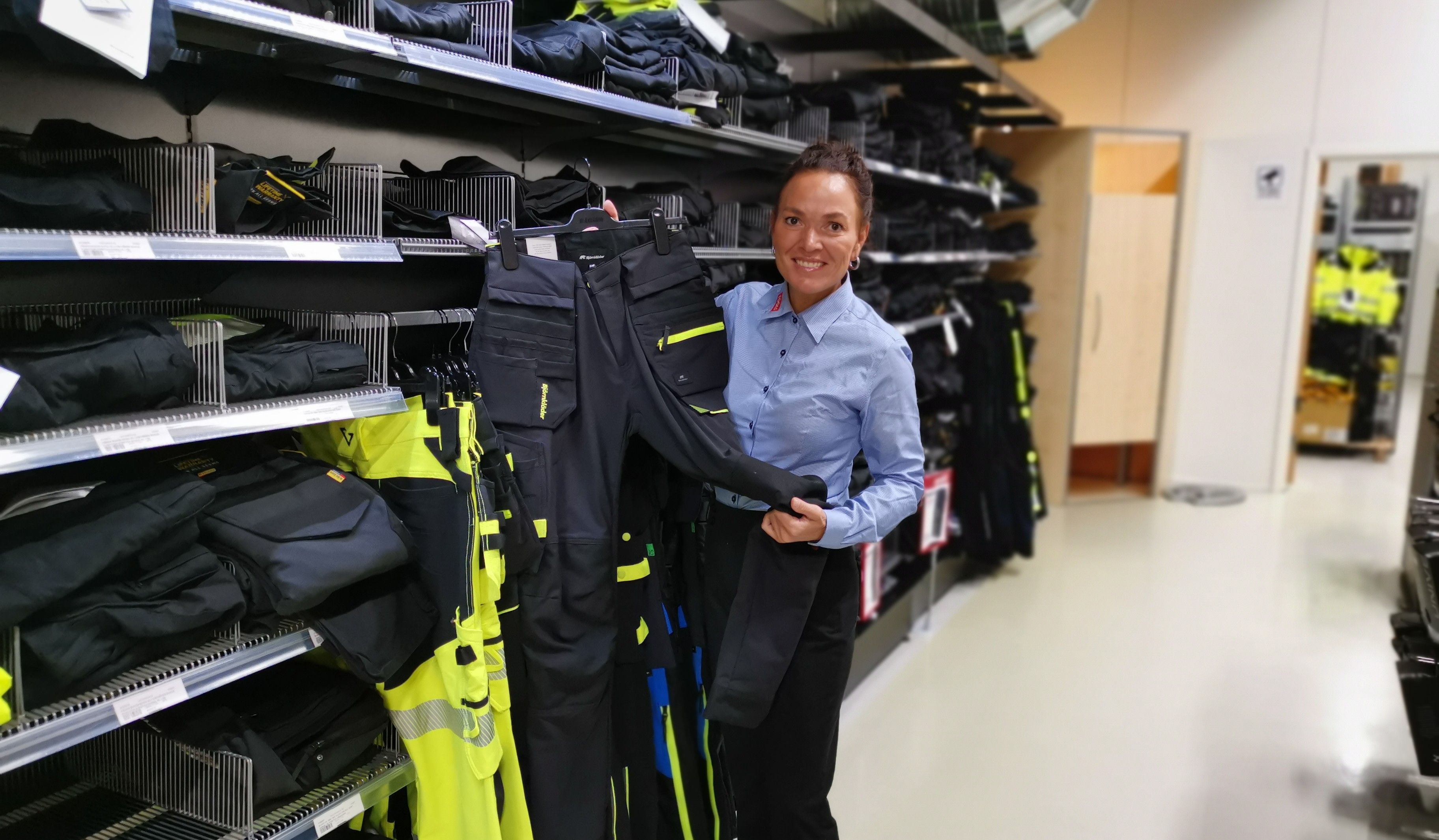 A person in a blue shirt holds work pants in a store aisle filled with various clothing items on shelves.