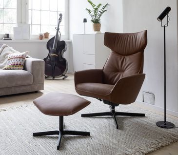 Conacfarbener Ledersessel mit passendem Fußhocker in hellem Wohnzimmer mit hellgrauem Sofa und weißem Highboard.