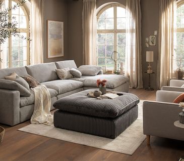 Wohnzimmer im gemütlichen Landhausstil mit beigem Ecksofa, zwei passenden Sesseln und einem dunkelgrauen Hocker.