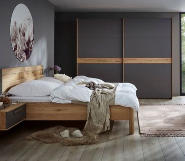 Grauer Hocker neben einem Massivholz Bettgestell mit weißer Bettwäsche.