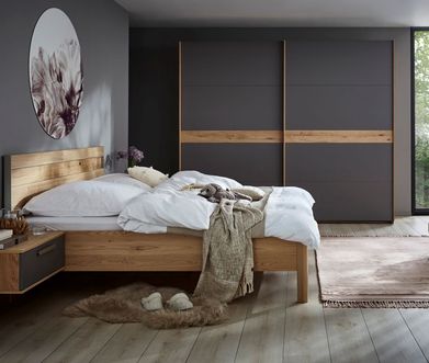 Grauer Hocker neben einem Massivholz Bettgestell mit weißer Bettwäsche.