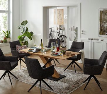 Heller Esstisch aus massivem Holz mit schwarzer Fußplatte neben schwarzen Polsterstühlen.