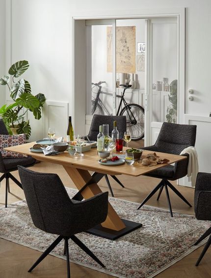 Heller Esstisch aus massivem Holz mit schwarzer Fußplatte neben schwarzen Polsterstühlen.