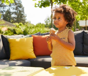Kleines Kind mit Eis vor Outdoor-Loungemöbeln mit gelben und orangen Kissen.
