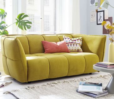 Viele Stoffsofas haben einen Bezug aus Mischgewebe, dass heißt aus Natur- sowie Kunstfasern Gelbes 2-Sitzer-Sofa vor großem Fenster mit bunten Kissen dekoriert.