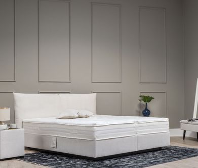 Beiges Boxspringbett neben einem farblich passenden Nachttisch mit einer modernen Lampe vor einer grauen Wand.