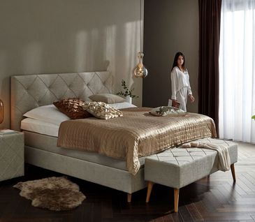 Schlafzimmer mit beigem Bett und goldener Tagesdecke. Sitzbank und Beistelltisch im gleichen Stil wie Bett.