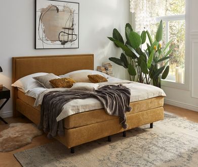 Gelbes Boxspringbett mit heller Bettwäsche und gelben Deko-Kissen.