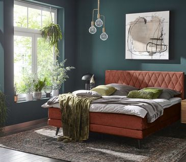 Boxspringbett in Kupferrot vor petrolfarbener Wand mit weißer und grüner Bettwäsche