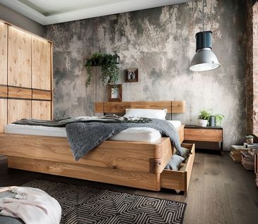 Helles Schlafzimmer mit Schrank und Bett aus Massivholz vor grauer Betonwand. Dekoriert mit grauer Tagesdecke und grünen Pflanzen auf Beistelltisch.