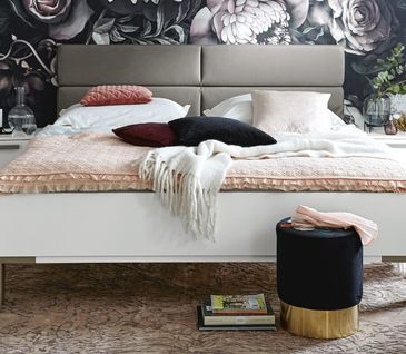 Weißes Bett mit hellgrauer Rückenlehne vor Wand mit auffälliger Blumentapete.