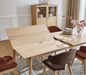 Verlängerbarer Esstisch im modernen Skandi-Design in hellem Massivholz umgeben von Esszimmerstühlen mit grauem und rotem Stoffbezug.