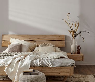 Massivholzbett mit heller Bettwäsche und Kissen dekoriert, mit Nachttisch auf dem eine Vase mit Blumen und andere Deko steht.