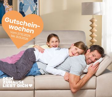 Vater liegt entspannt mit zwei jungen Töchtern auf gemütlichem Sofa in hellem Wohnzimmer.