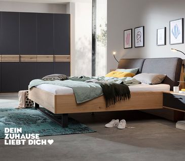 Drehtürenschrank mit dunkelgrauer Front und hellen Holzakzenten und modernem Bett mit gepolstertem Kopfteil.