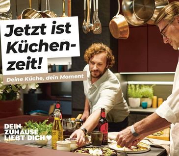 Zwei Männer kochen in moderner Küche mit roten Fronten. Töpfe hängen von oben herab.