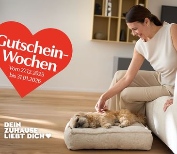 Elegant gekleidete Frau sitzt auf weißem Sofa uns streichelt Hund, der auf passendem Hundebett liegt.