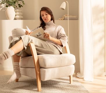 Frau sitzt in bequemen Sessel mit beigem Bouclé-Stoff und liest gemütlich in minimalistisch eingerichteten Wohnzimmer.