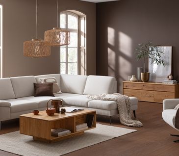 Wohnzimmer im Skandi-Stil mit weißem Ecksofa, passendem Relaxsessel, Couchtisch aus Holz und braun gestrichener Wand.
